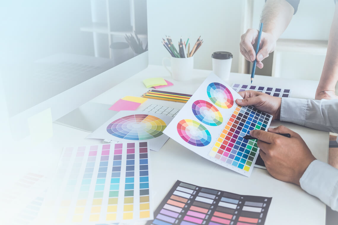Două persoane colaborează la un birou cu mostre de culoare, roți de culoare și un computer. Unul indică o diagramă de culori în timp ce celălalt o ține. Biroul este împrăștiat cu diagrame, note lipicioase, o ceașcă de cafea și rechizite de artă, creând un mediu care sugerează un spațiu de lucru creativ concentrat pe design și strategii SEO.